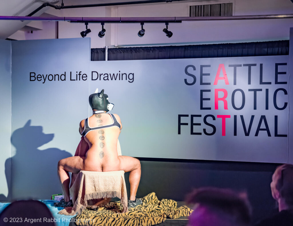 2025 Gallery Hours: The 2024 SEAF Life Drawing Stage. A man sitting backwards on a chair, with his back towards the camera, wearing a harness and a dog mask. A small crowd is around him, drawing.