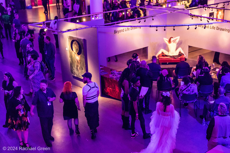 SEAF 2025 Saturday: A picture taken from above of the 2024 Seattle Erotic Art Festival. The image focuses on crowds around the Art Studio area while a model poses on a couch for several people drawing her and other guests walk past.